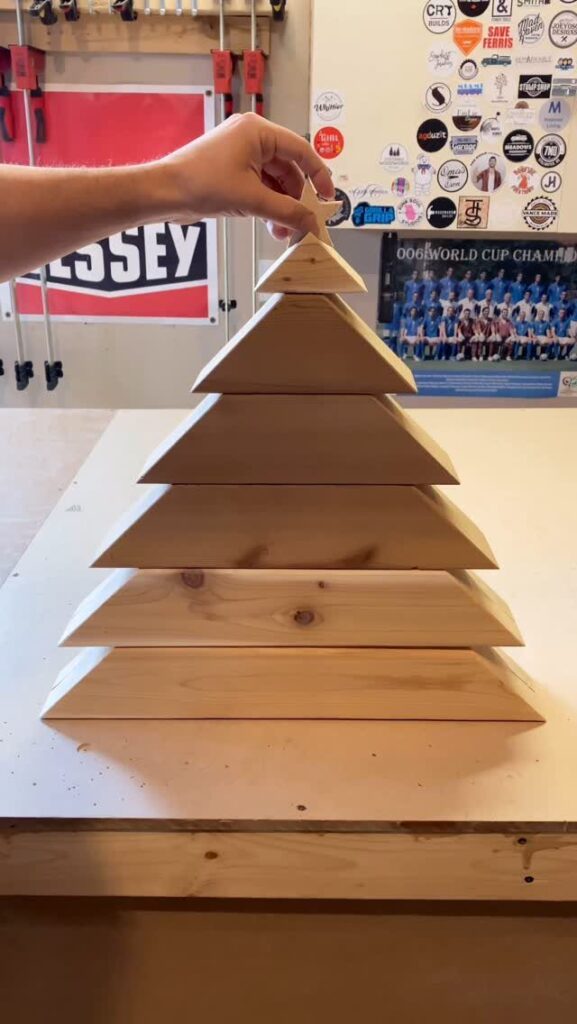 A hand placing a wooden star atop a tiered pine wood Christmas tree on a workbench.