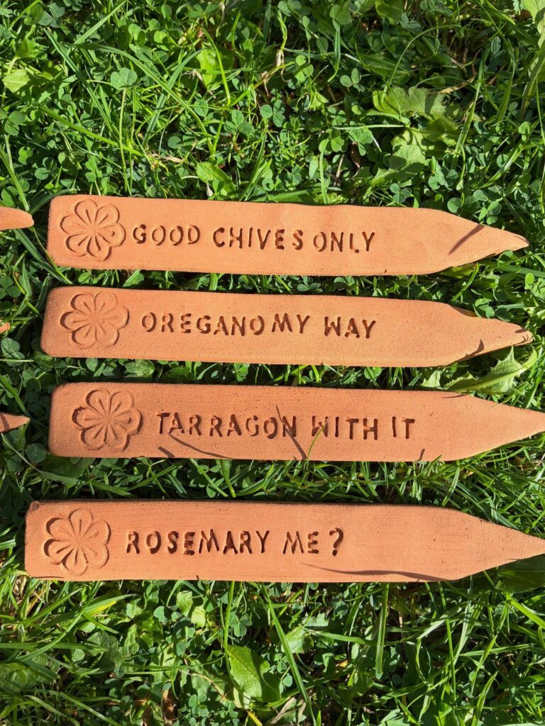Terracotta herb garden signs with puns lying on green grass.