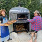 Two people using a large mosaic-tiled outdoor pizza oven to cook and serve fresh pizzas.