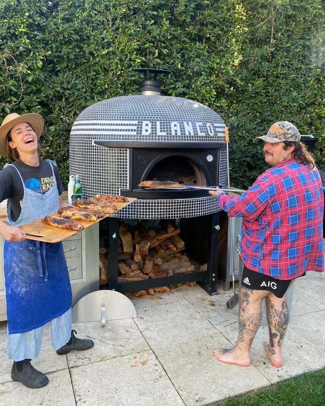 Two people using a large mosaic-tiled outdoor pizza oven to cook and serve fresh pizzas.