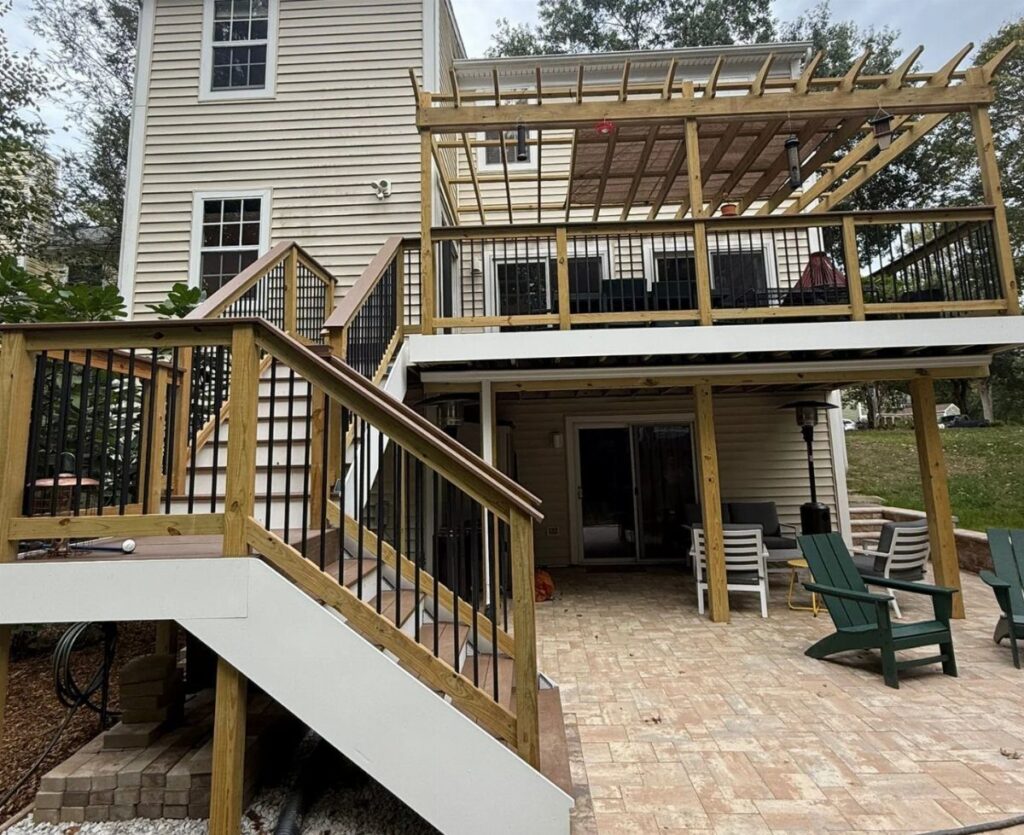 Wooden elevated deck with black railings overlooking a paved stone patio with outdoor furniture underneath.