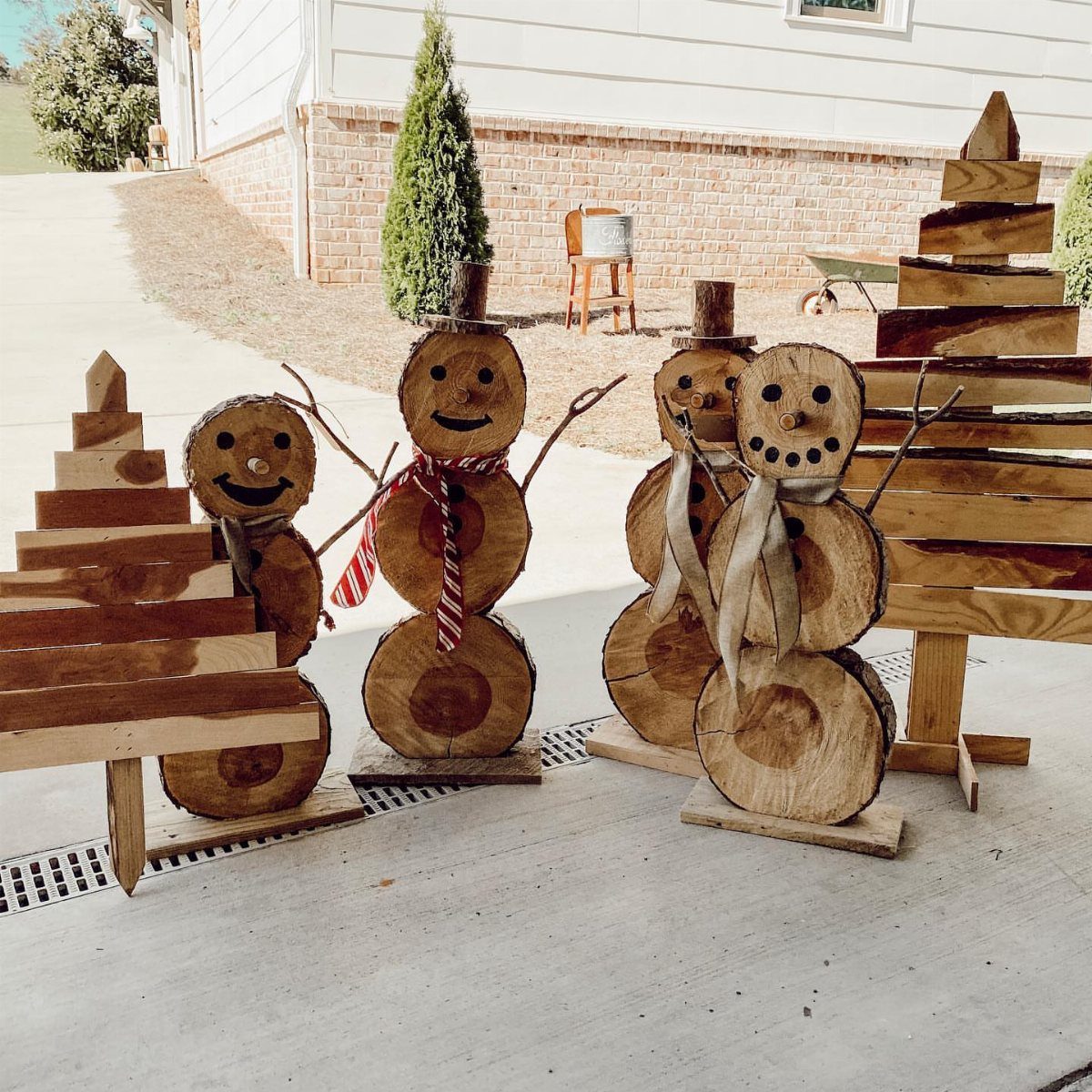 Four rustic wooden Christmas decorations, including two snowmen and two trees, sitting on a concrete patio.